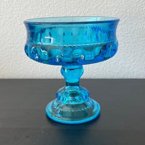 May include: A vintage, turquoise-colored glass pedestal bowl. The bowl has a wide, rounded top with a decorative pattern around the rim. The stem and base are also turquoise and feature similar detailing. The bowl is likely used for serving desserts or displaying small items.