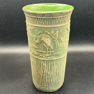 May include: A cylindrical green vase featuring a textured surface with a relief design of a bird standing in a marshy landscape. The upper and lower sections have vertical ribbed patterns, while the rim is smooth and finished in a lighter green.
