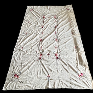 May include: Cream-colored tablecloth with intricate floral embroidery in shades of red, pink, and blue. The embroidery features a central vertical design and corner accents, creating a vintage aesthetic. The tablecloth is rectangular and appears to be made of a soft fabric.