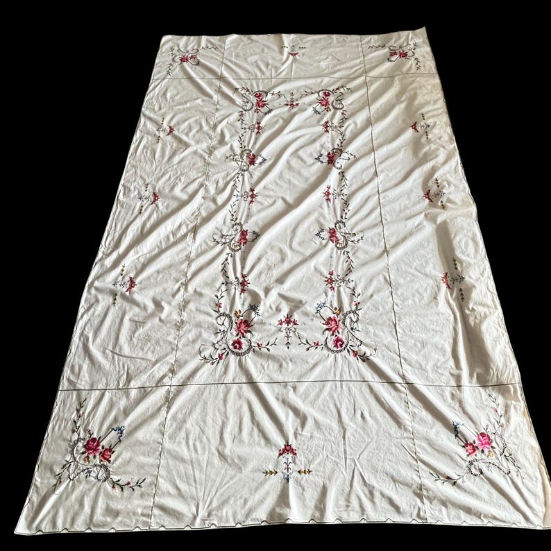 May include: Cream-colored tablecloth with intricate floral embroidery in shades of red, pink, and blue. The embroidery features a central vertical design and corner accents, creating a vintage aesthetic. The tablecloth is rectangular and appears to be made of a soft fabric.