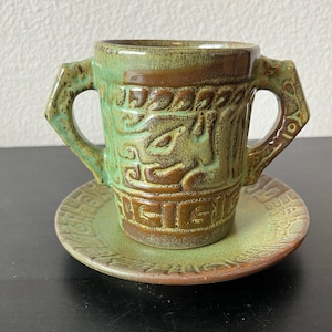 Op de afbeelding: Een keramische kop en schotel set met een groene en bruine glazuur. De kop heeft twee handvatten en is versierd met reliëfpatronen. De schotel heeft ook reliëf ontwerpen. De set is waarschijnlijk handgemaakt.