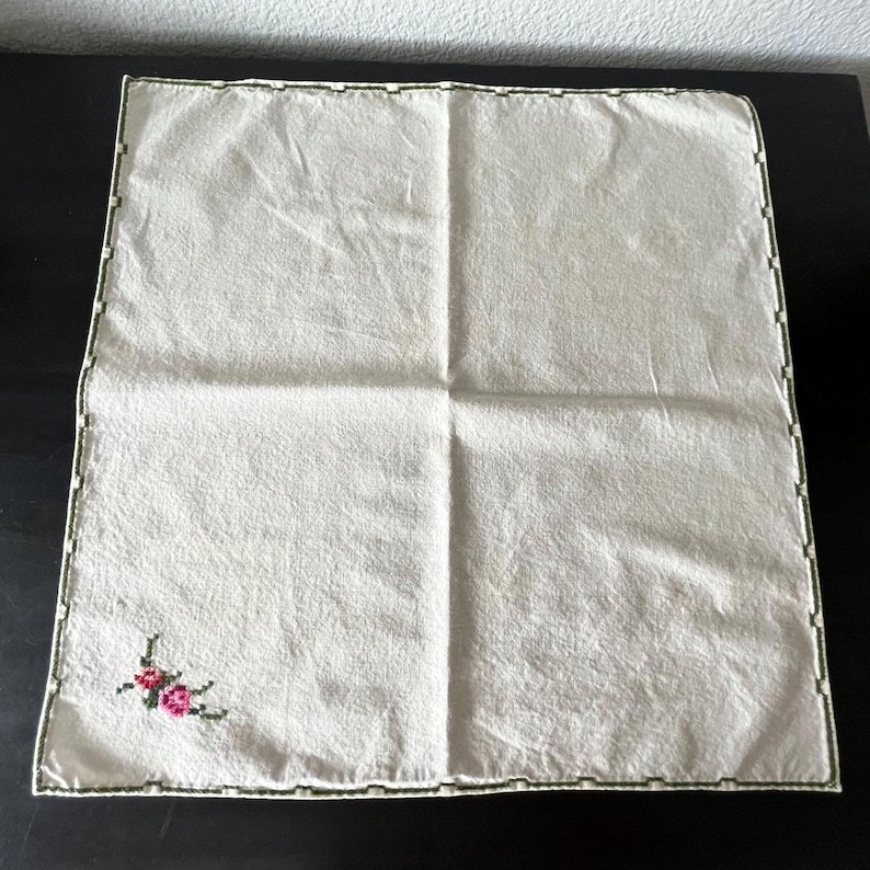 May include: Off-white square handkerchief with a green border and a small floral embroidery in the corner. The embroidery features pink and red flowers with green leaves. The handkerchief is folded and lies flat on a dark surface.