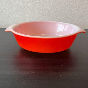 May include: A round, shallow baking dish with two small side handles. The exterior of the bowl is a vibrant, solid orange color, while the interior features a smooth, pale white finish. The dish has a wide, open top and a curved base.