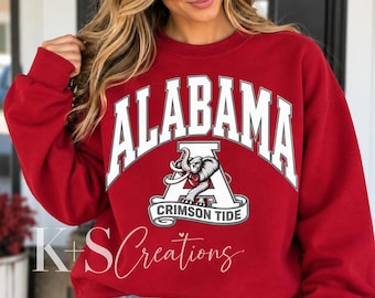 May include: A crimson red crewneck sweatshirt featuring the word ALABAMA in large white arched letters above a white letter A logo with a grey elephant graphic and the text CRIMSON TIDE on a banner. The sweatshirt has long sleeves and a ribbed collar.