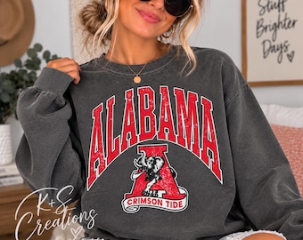 May include: A charcoal grey crewneck sweatshirt featuring a distressed red graphic with the word ALABAMA in an arched font above a large letter A containing an elephant illustration and the text CRIMSON TIDE on a white banner at the bottom.