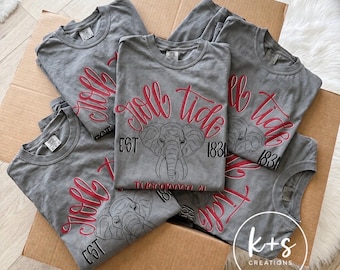 May include: A collection of grey t-shirts featuring a red script graphic that reads Roll Tide, a black line-art elephant illustration, the text EST 1831, and the word Tuscaloosa, AL in red. The shirts are folded and arranged inside a cardboard box.