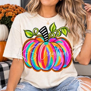 May include: A cream-coloured t-shirt featuring a large, vibrant graphic of a pumpkin. The pumpkin is decorated with colourful, abstract brushstrokes in shades of bright pink, orange, blue, and yellow, topped with green leaves and a black and white striped stem.