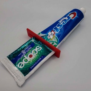 May include: A red plastic tube squeezer attached to a tube of Crest Plus Scope Whitening Outlast toothpaste. The toothpaste tube is white with green and blue accents and the text "Scope Whitening Outlast" is printed on the tube.