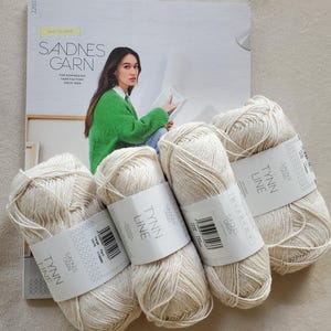 May include: Five skeins of off-white yarn labeled "TYNN LINE" and "SANDNES GARN" are arranged in front of a magazine. The magazine features a woman wearing a green sweater. The text "MYKT TIL DAME" is visible.