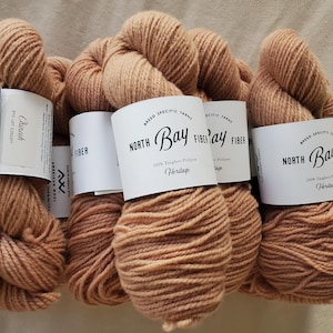 May include: Several skeins of tan-colored yarn are bundled together. Each skein has a white label with black text that reads "North Bay Fiber" and "Heritage." One label reads "Chinook." The yarn is made of Targhee-Polypay fiber.