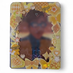 May include: A rectangular plastic card holder decorated with yellow and orange stickers featuring hearts, stars, butterflies, a dog, and the word sweet. The border is layered with glittery and holographic adhesive shapes surrounding a central blurred image.