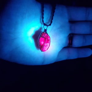 May include: A pink crystal pendant on a black cord necklace. The crystal is wrapped in wire and glows under a blue light.