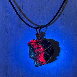 May include: A pendant featuring a raw, irregularly shaped stone wrapped in dark metal wire, suspended from a simple black cord. The stone exhibits a vibrant, glowing red and dark green surface under ultraviolet light, creating a high-contrast effect.