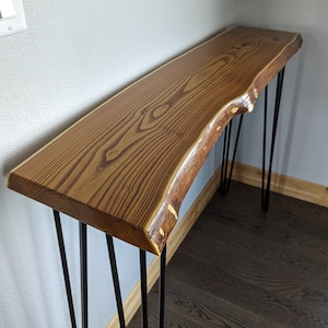 May include: A wooden console table with a live edge top and black hairpin legs. The table has a natural wood finish and a rustic look.