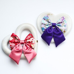 May include: Two fabric bow ties, one with a red and white floral pattern and a pink satin back, and the other with a purple satin back and a blue and white floral pattern.