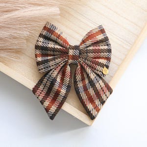 May include: A brown, black, and beige plaid bow with a gold accent. The bow is tied in a classic style and is perfect for adding a touch of whimsy to any outfit.