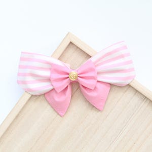 May include: A pink and white striped bow with a gold button in the center. The bow is on a wooden surface.