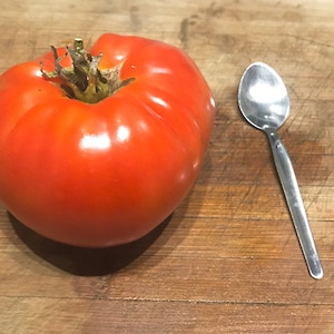 May include: A large, bright red tomato next to a silver teaspoon on a wooden cutting board.