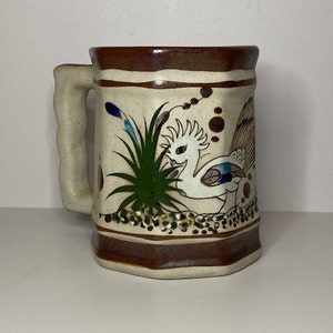 May include: A brown and white ceramic mug with a handle. The mug is decorated with a white bird with blue and green accents, surrounded by green leaves and brown dots.