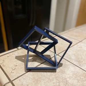 Könnte beinhalten: Eine blaue geometrische Skulptur aus drei ineinandergreifenden Quadraten. Die Skulptur ist aus dunkelblauem Material gefertigt und hat ein modernes, abstraktes Design.