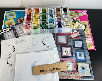 May include: A clear plastic organiser box filled with colourful embroidery floss bobbins, cross-stitch pattern leaflets, white fabric with blue printed designs, and a wooden ruler labelled Nanny Goat Notions measuring 15 cm in length.