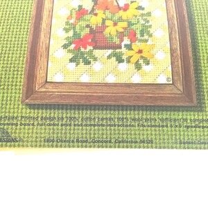 May include: A needlepoint kit package featuring a floral basket design in a brown frame on a green background. Text includes: This kit contains: Printed design on 100% cotton canvas, 100% wool yarns, needle, mounting board, full colour print and instructions.