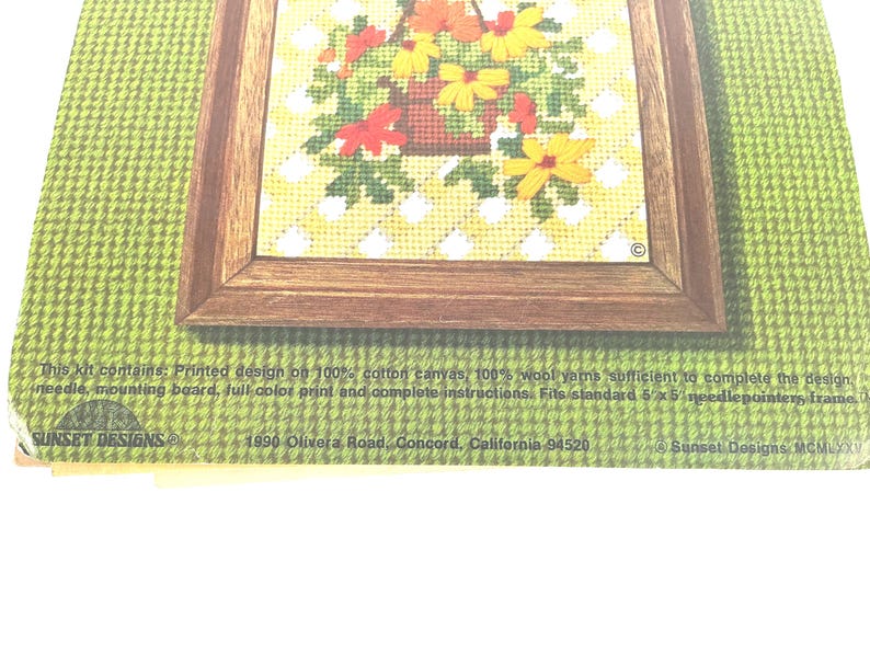May include: A needlepoint kit package featuring a floral basket design in a brown frame on a green background. Text includes: This kit contains: Printed design on 100% cotton canvas, 100% wool yarns, needle, mounting board, full colour print and instructions.