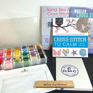 May include: A clear plastic organizer box filled with multi-colored embroidery floss bobbins, three cross-stitch pattern books, a white DMC branded folder, and a wooden ruler labeled Nanny Goat Notions measuring 6 inches in length.