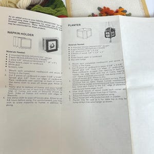 May include: A white instructional paper sheet for a needlepoint kit featuring text and diagrams for a napkin holder and a planter. The paper includes lists of materials needed and numbered assembly steps. Bundles of colourful yarn are visible nearby.
