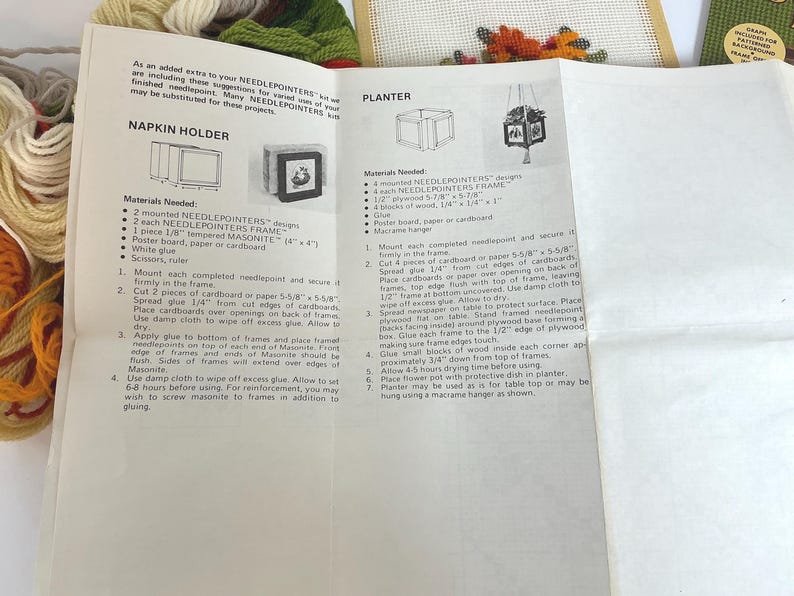 May include: A white instructional paper sheet for a needlepoint kit featuring text and diagrams for a napkin holder and a planter. The paper includes lists of materials needed and numbered assembly steps. Bundles of colourful yarn are visible nearby.