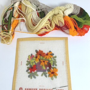 May include: A needlepoint canvas featuring a floral design with orange, yellow, and green threads, labelled Sunset Designs 1973. The kit includes a bundle of assorted yarn in shades of cream, yellow, orange, brown, and green for embroidery.