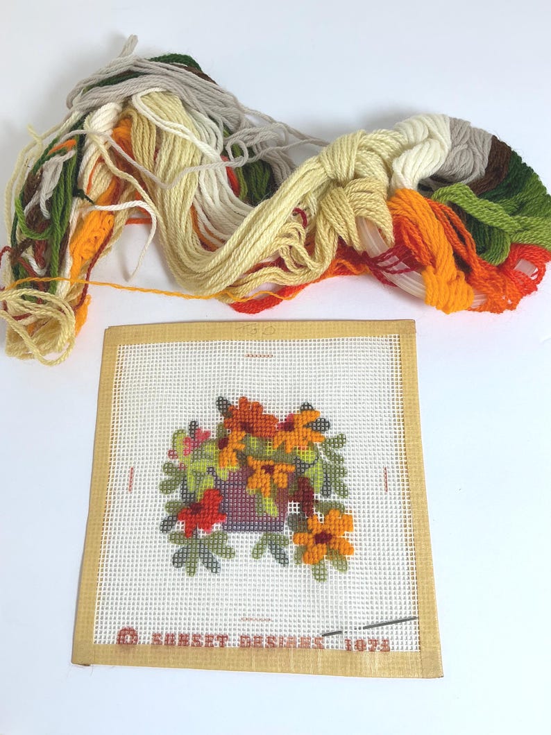 May include: A needlepoint canvas featuring a floral design with orange, yellow, and green threads, labelled Sunset Designs 1973. The kit includes a bundle of assorted yarn in shades of cream, yellow, orange, brown, and green for embroidery.