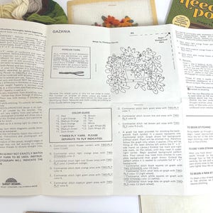 May include: A white instructional paper for a Gazania needlepoint kit by Sunset Designs, featuring a black and white line drawing of flowers in a pot, a colour guide, and detailed stitching instructions for the project, with yarn bundles visible.