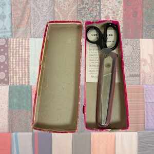 May include: Vintage sewing scissors in a red and pink box. The scissors are black with a silver handle and the text "JOYING" is visible. The box is open, revealing the scissors and a paper insert with text. The background is a collage of fabric patterns.