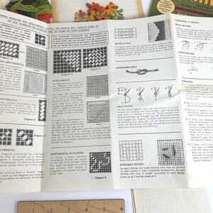 May include: A white instructional needlepoint guide featuring black text and diagrams for various stitches, including horizontal continental, basketweave, and satin stitch. A wooden ruler with metric markings is visible at the bottom edge of the paper.
