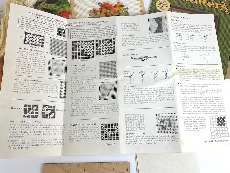 May include: A white instructional needlepoint guide featuring black text and diagrams for various stitches, including horizontal continental, basketweave, and satin stitch. A wooden ruler with metric markings is visible at the bottom edge of the paper.