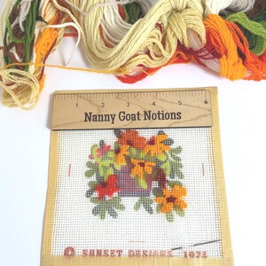 May include: A needlepoint canvas featuring a floral arrangement in a purple pot, with a wooden ruler labelled Nanny Goat Notions measuring 15 cm across the top. A bundle of embroidery floss in green, yellow, orange, and white sits above the canvas.