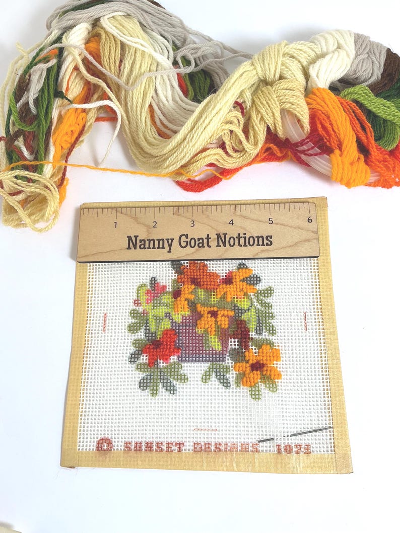 May include: A needlepoint canvas featuring a floral arrangement in a purple pot, with a wooden ruler labelled Nanny Goat Notions measuring 15 cm across the top. A bundle of embroidery floss in green, yellow, orange, and white sits above the canvas.