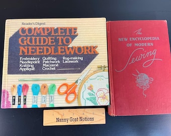 May include: A Reader's Digest Complete Guide to Needlework book and a red New Encyclopedia of Modern Sewing book. A wooden ruler labelled Nanny Goat Notions is placed below the books. The guide features embroidery floss, scissors, and a needle.