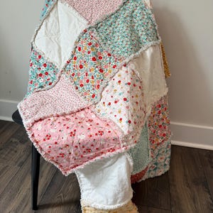 May include: A patchwork rag quilt featuring a variety of floral and gingham fabric squares in shades of pink, white, teal, and yellow. The quilt has exposed frayed seams between each square and a small rectangular label at the bottom corner.