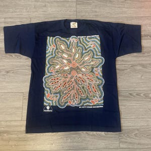 May include: Dark blue t-shirt with a detailed, multi-coloured pattern in orange, white, and blue. The shirt displays the text "WITCHETTY GRUB DREAMING" and "BY: KITTY EVANS NAKAMARRA". The garment is laid flat on a light wooden surface.