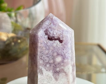 May include: A lavender-colored crystal tower with a geode-like cavity at the top. The crystal has a hexagonal base and a pointed top. The surface has a textured, mottled appearance with white and purple hues. The crystal is displayed on a white marble surface.