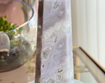 May include: A light purple crystal obelisk, standing on a white marble base. The obelisk has a pointed top and a rectangular shape, with a mix of light and dark purple hues. The crystal is a decorative item.