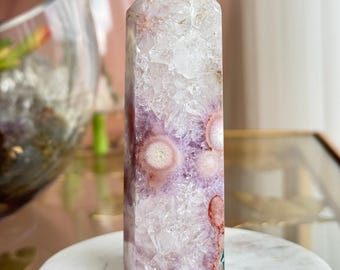 May include: A polished crystal tower featuring a hexagonal point, composed of translucent white and pale purple quartz with distinct circular pink and white flower-like inclusions scattered across the faceted, crystalline surface.