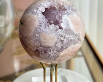 May include: A spherical amethyst geode featuring a polished, pale pink and lavender exterior with a central opening revealing jagged, translucent purple crystal formations. The geode is supported by a three-pronged gold metal stand on a clear base.
