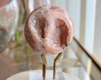 May include: A polished pink sphere with a central geode cavity filled with small, sparkling crystals. The sphere is supported by a gold-toned metal tripod stand attached to a clear, square acrylic base, creating a display for the mineral specimen.