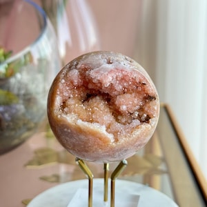 May include: A spherical geode, peach-colored on the exterior, with a cavity filled with sparkling pink crystals. The geode is displayed on a gold-colored metal stand with a white marble base, showcasing its natural beauty.