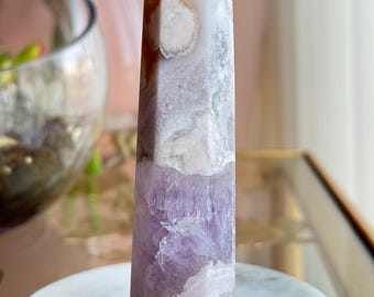 May include: A polished crystal tower featuring a hexagonal point, with a base of translucent light purple amethyst transitioning into a textured upper section of white and cream-colored agate with brown inclusions and a distinct circular pattern.