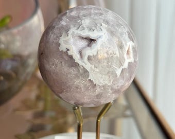 May include: A spherical, light purple crystal geode with white and light purple interior details. The geode is displayed on a gold-colored metal stand with a clear acrylic base, set on a white marble surface.
