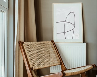 Puede incluir: Una silla de mimbre marrón claro con un marco de madera se encuentra en una habitación con un radiador blanco y una impresión de arte abstracto enmarcada con un diseño de línea púrpura sobre un fondo gris claro.